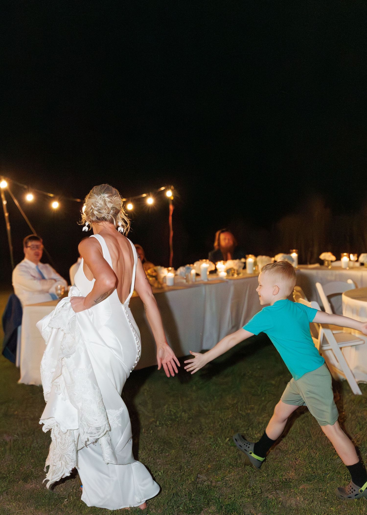 Bride in white gown dances with young boy under string lights at outdoor evening wedding reception.