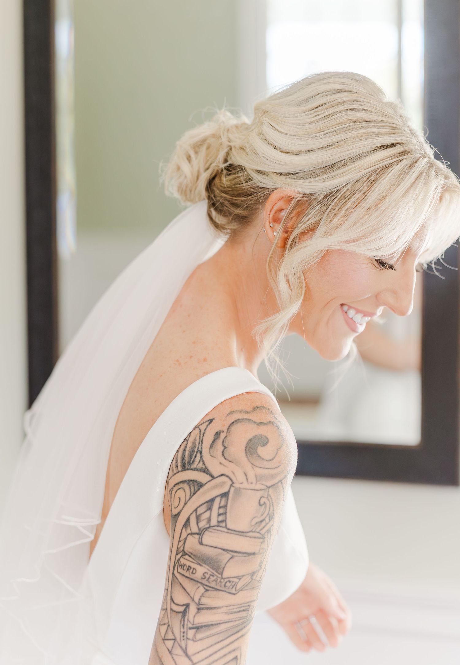 Smiling blonde bride with sleeve tattoo wearing updo hairstyle and veil on her wedding day.