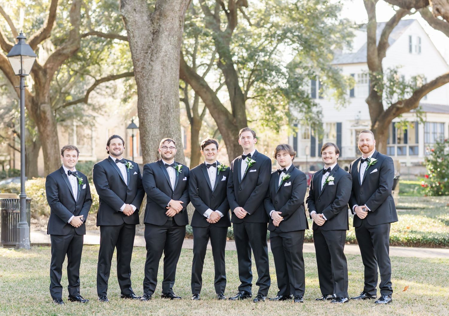 Groom and seven groomsmen in black tuxedos posing outdoors under large oak trees.