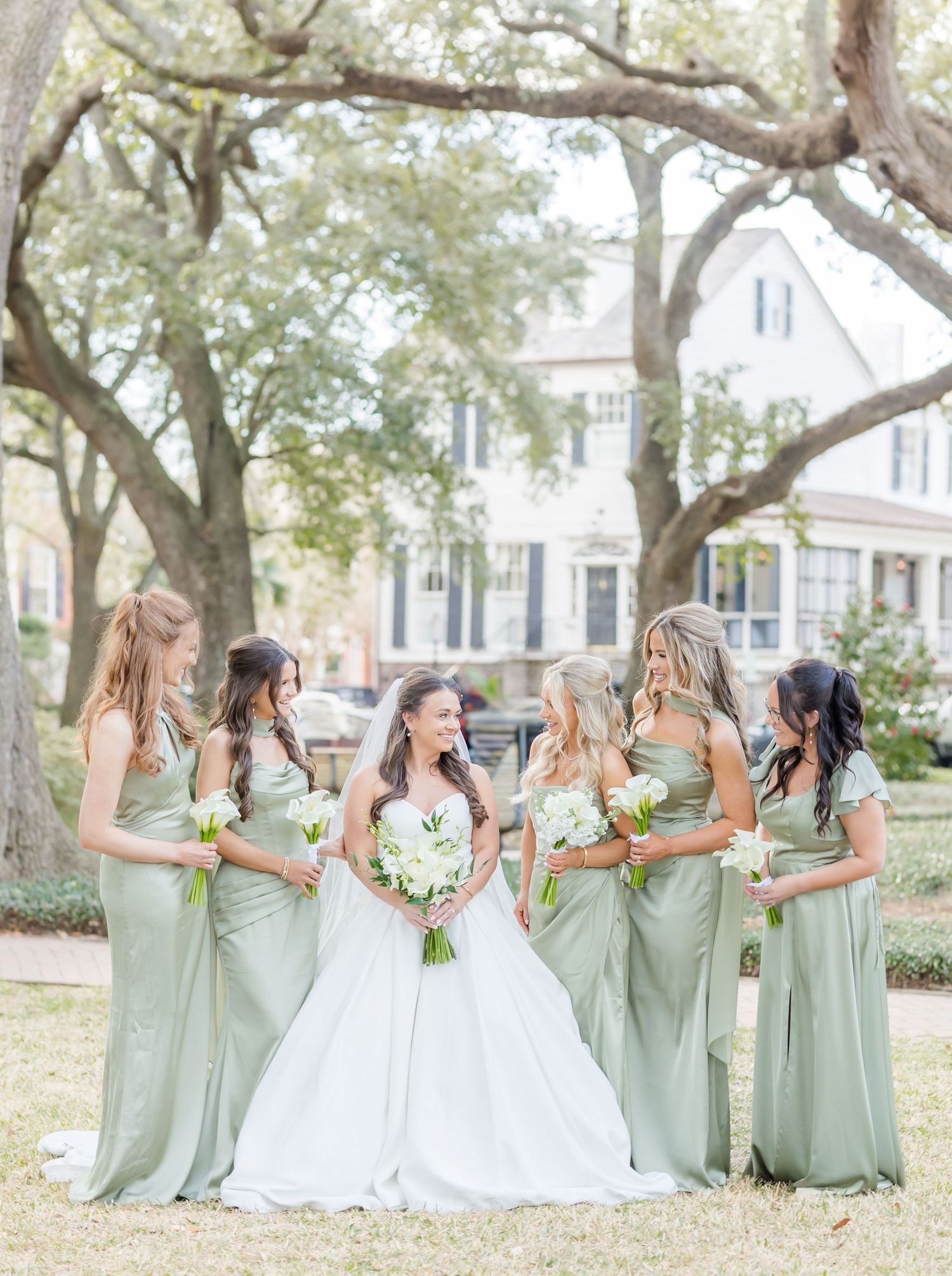 Bride in white gown with five bridesmaids in sage green dresses holding white bouquets outdoors.