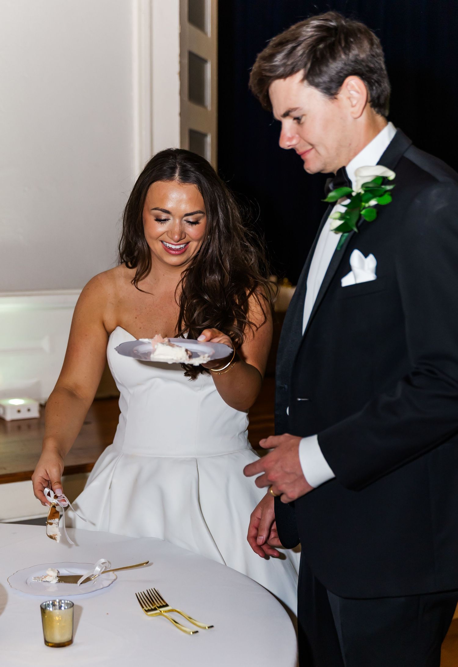Smiling bride in strapless gown holds cake plate while groom in tuxedo looks on at reception.