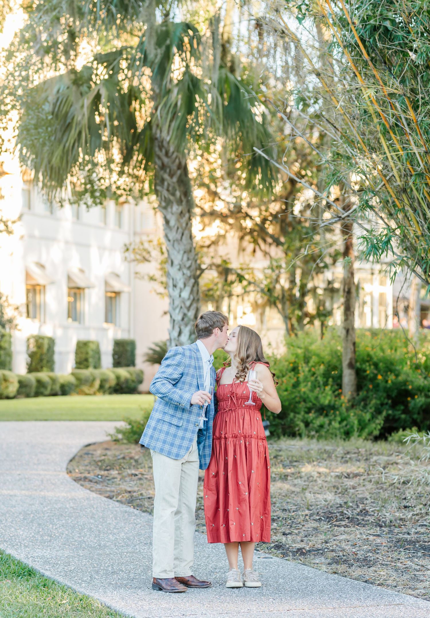 A couple in blue and coral attire share romantic moments together on a palm tree-lined path at sunset.