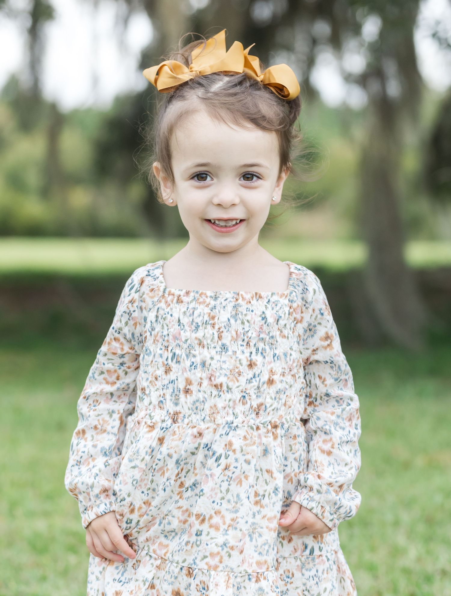 Young child in floral dress and yellow hair bow posing on grass.