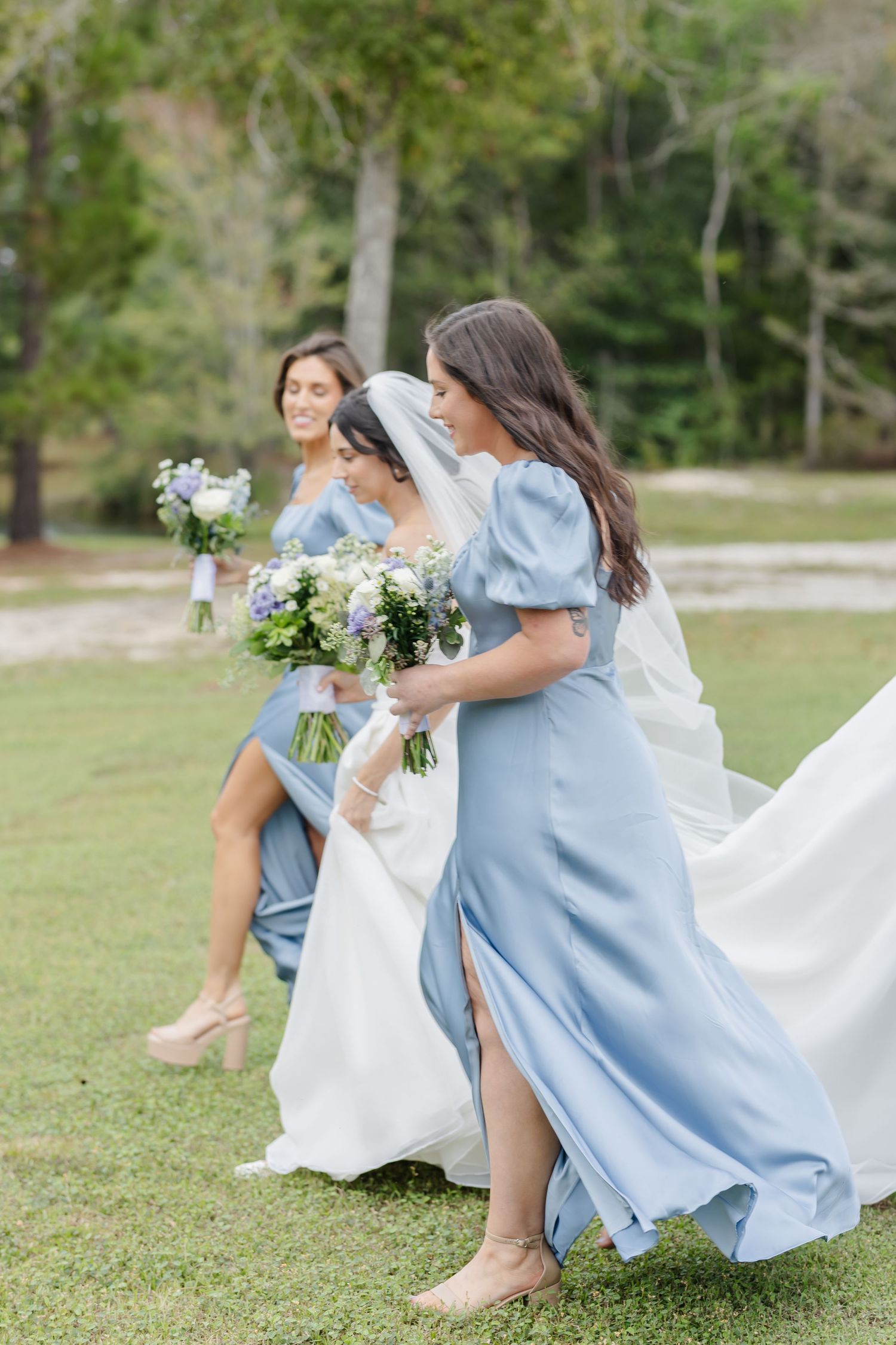 Bridesmaids in powder blue dresses help with bride's flowing train outdoors.