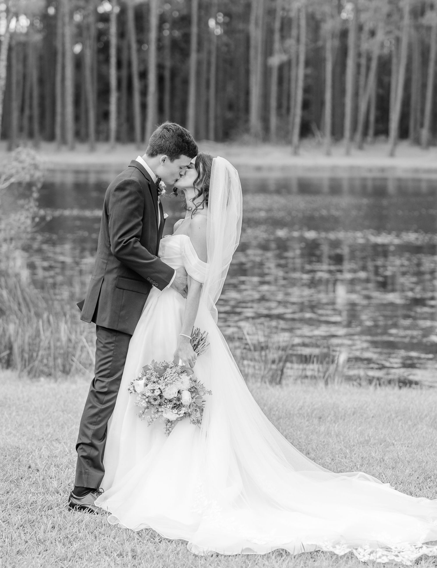 A romantic black and white portrait of a wedding couple embracing by a lake.