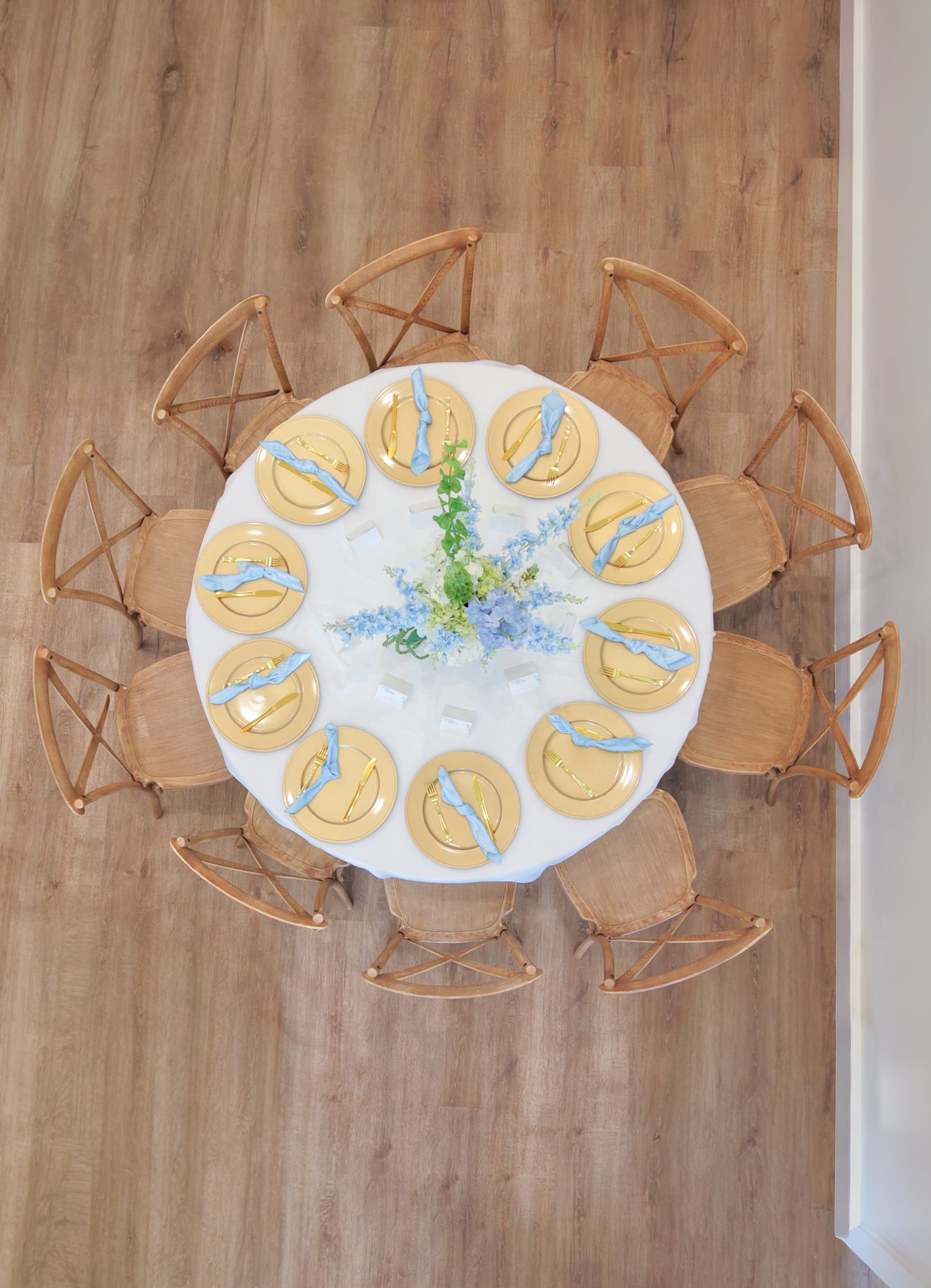 Aerial view of a round white dining table with gold plates, blue napkins, and floral centerpiece, surrounded by wooden chairs.