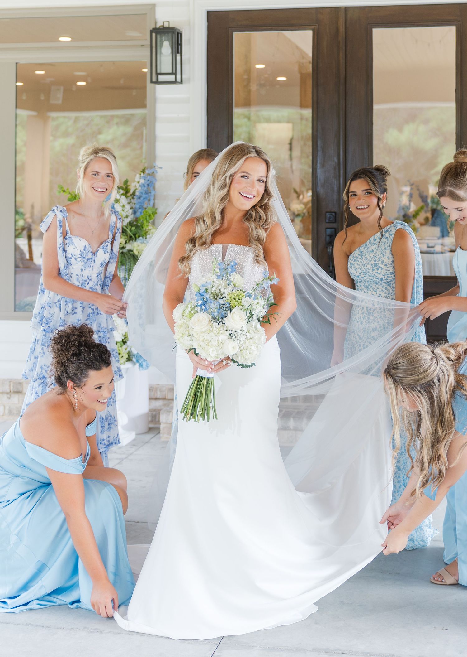 Bride in white gown holds blue and white bouquet while bridesmaids in blue dresses arrange her veil outdoors.