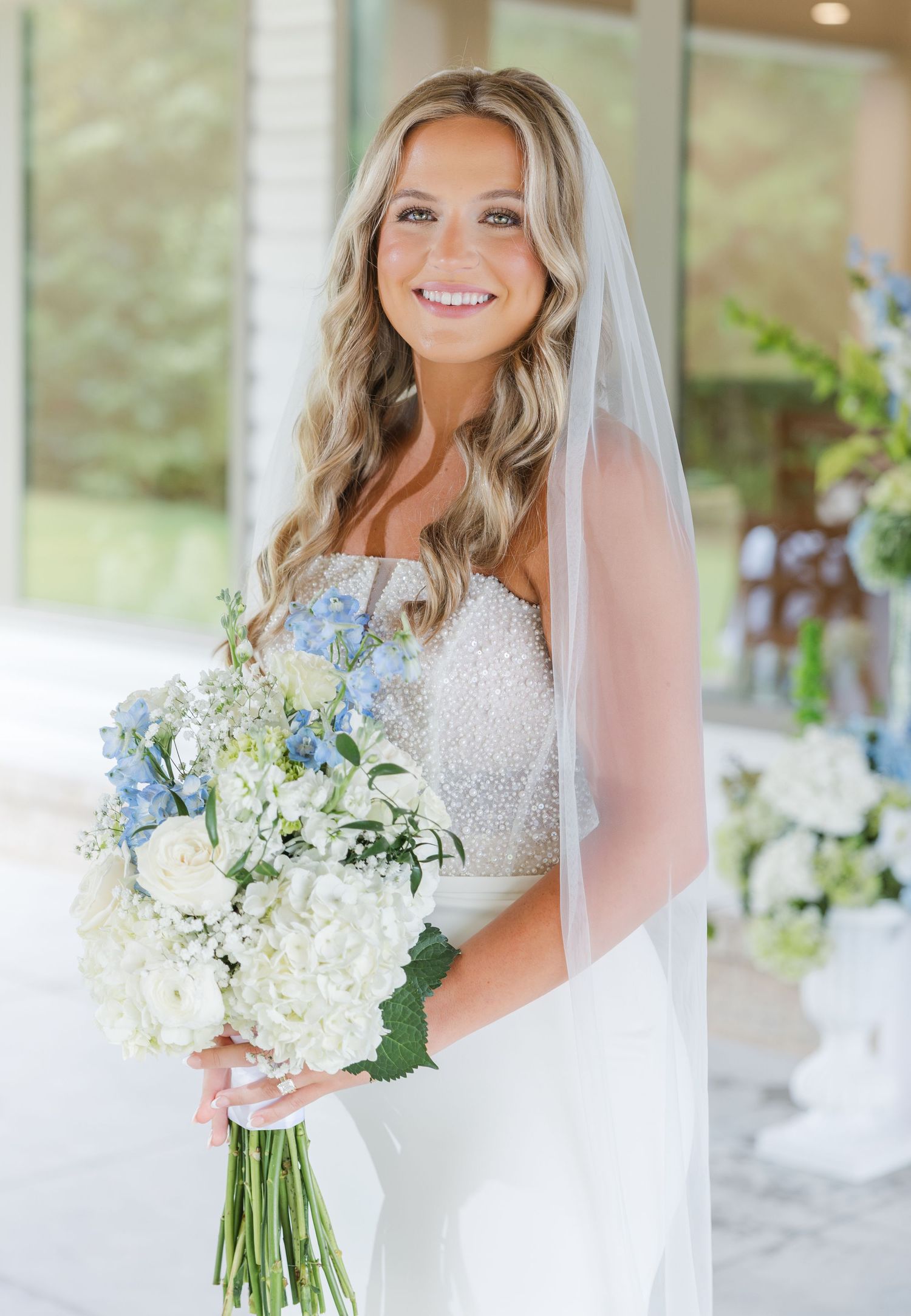 Beautiful blonde bride in beaded strapless gown holding blue and white hydrangea bouquet with veil.
