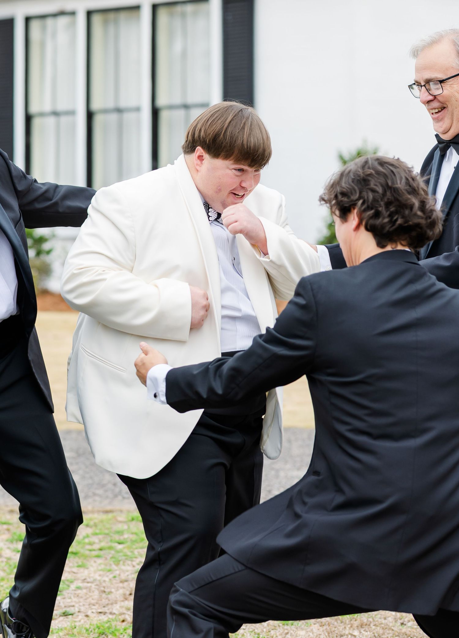 Groom in ivory tuxedo playfully sparring with groomsmen in black suits on the lawn during wedding day celebrations.