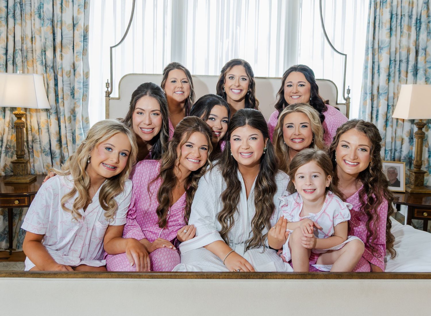 Bride and bridesmaids in pink robes smiling together on a bed during wedding morning preparations.