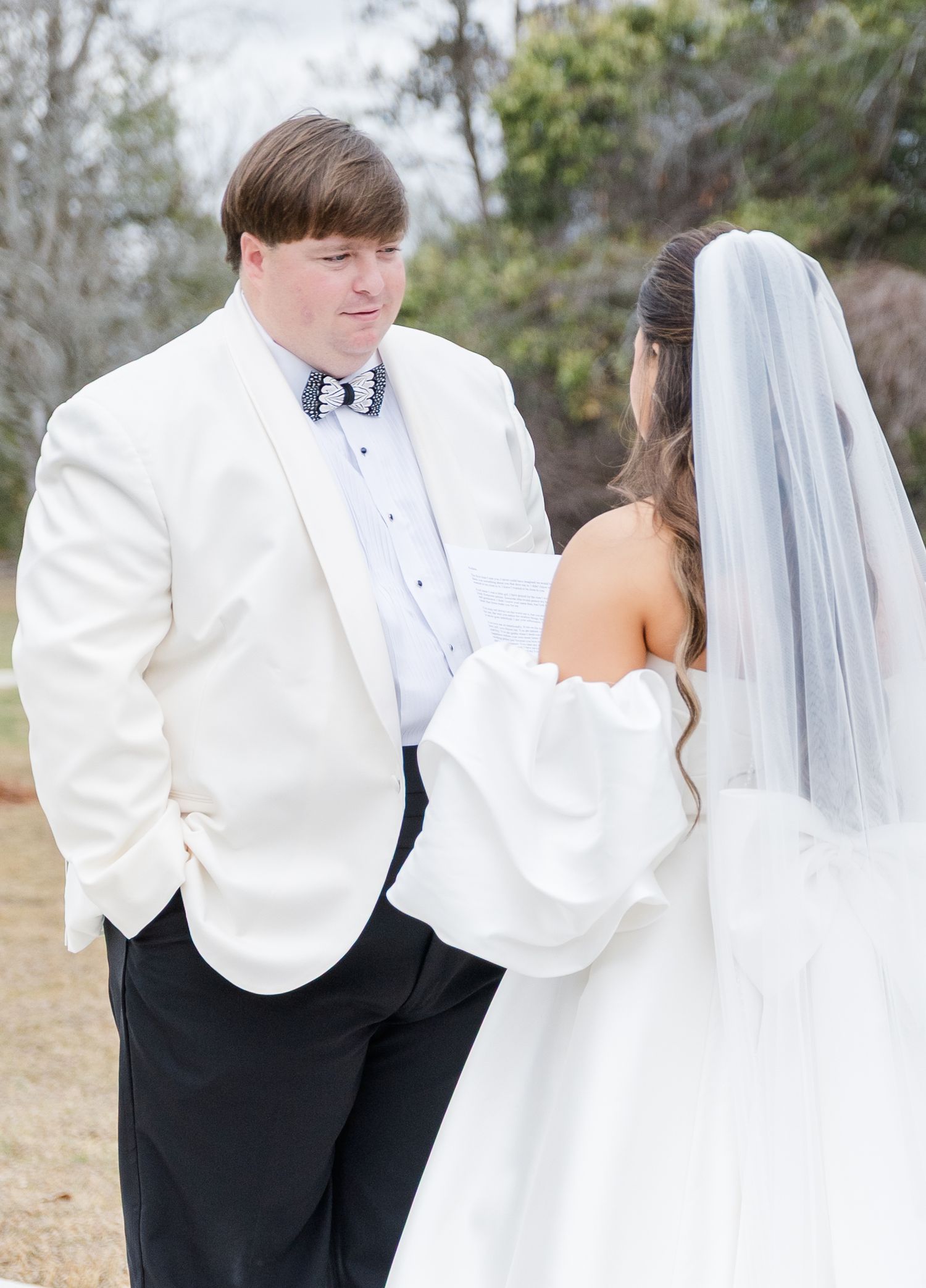 Groom in white tuxedo gazes at bride in strapless satin gown during outdoor first look moment.