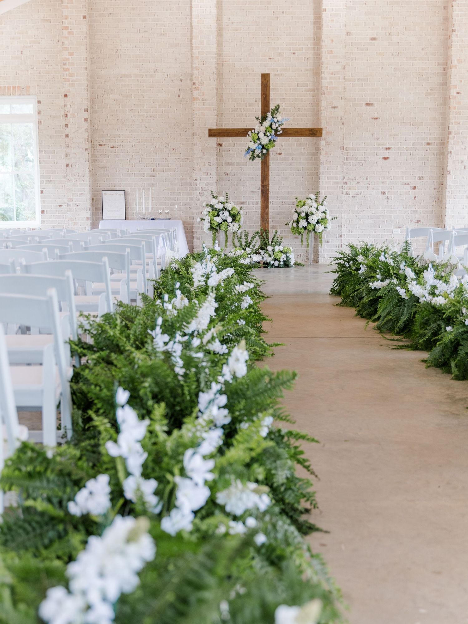 Rustic wedding ceremony venue with white chairs, lush green fern aisle runners, white flowers, and a wooden cross altar.