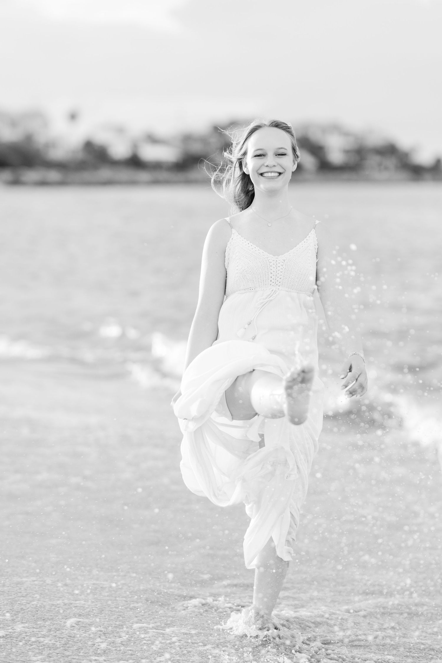 A joyful figure in a white sundress splashes in shallow beach water on a sunny day, captured in black and white.