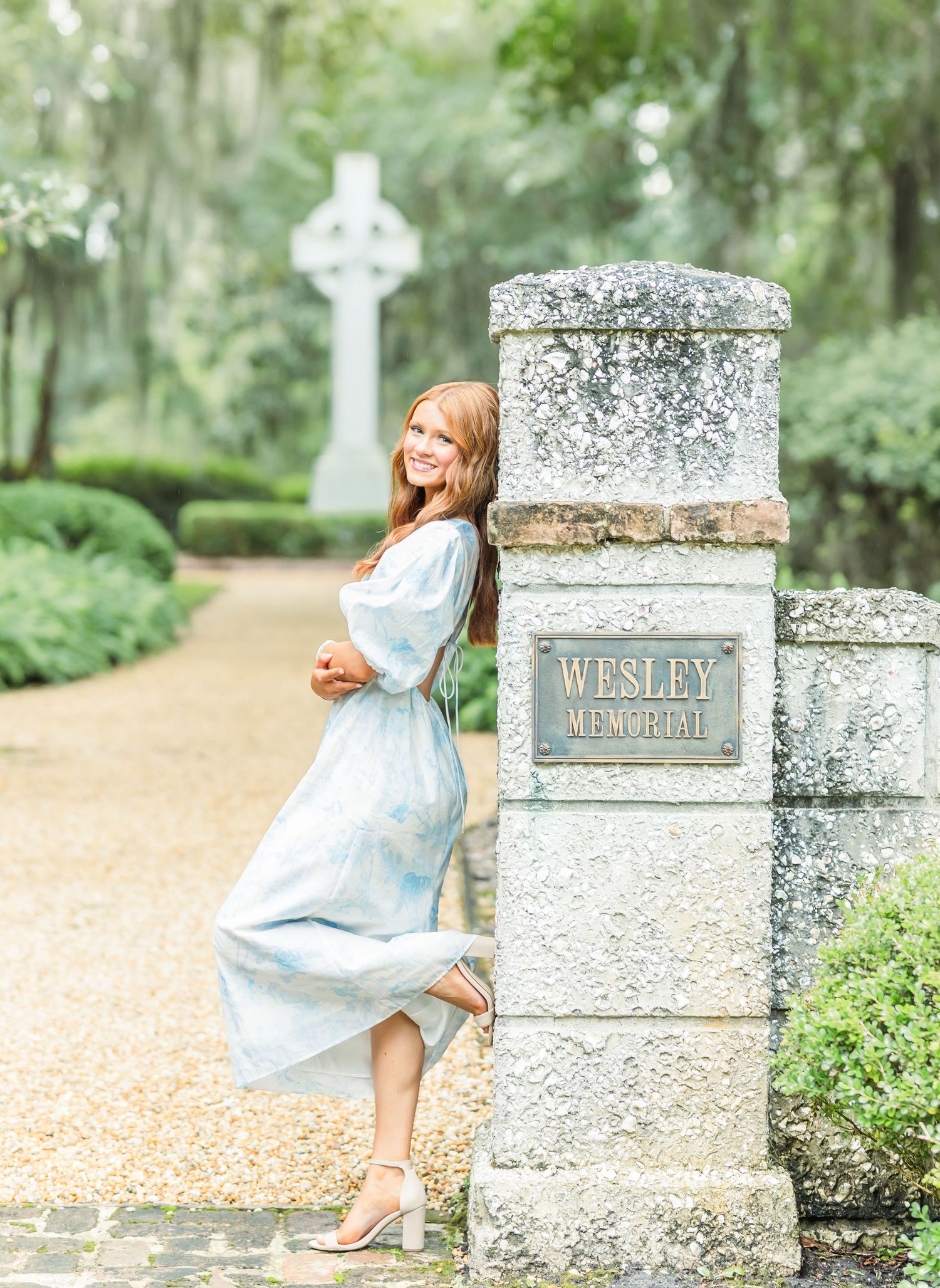 A person in a light blue dress poses gracefully near old stone pillars along a pathway in a garden setting.