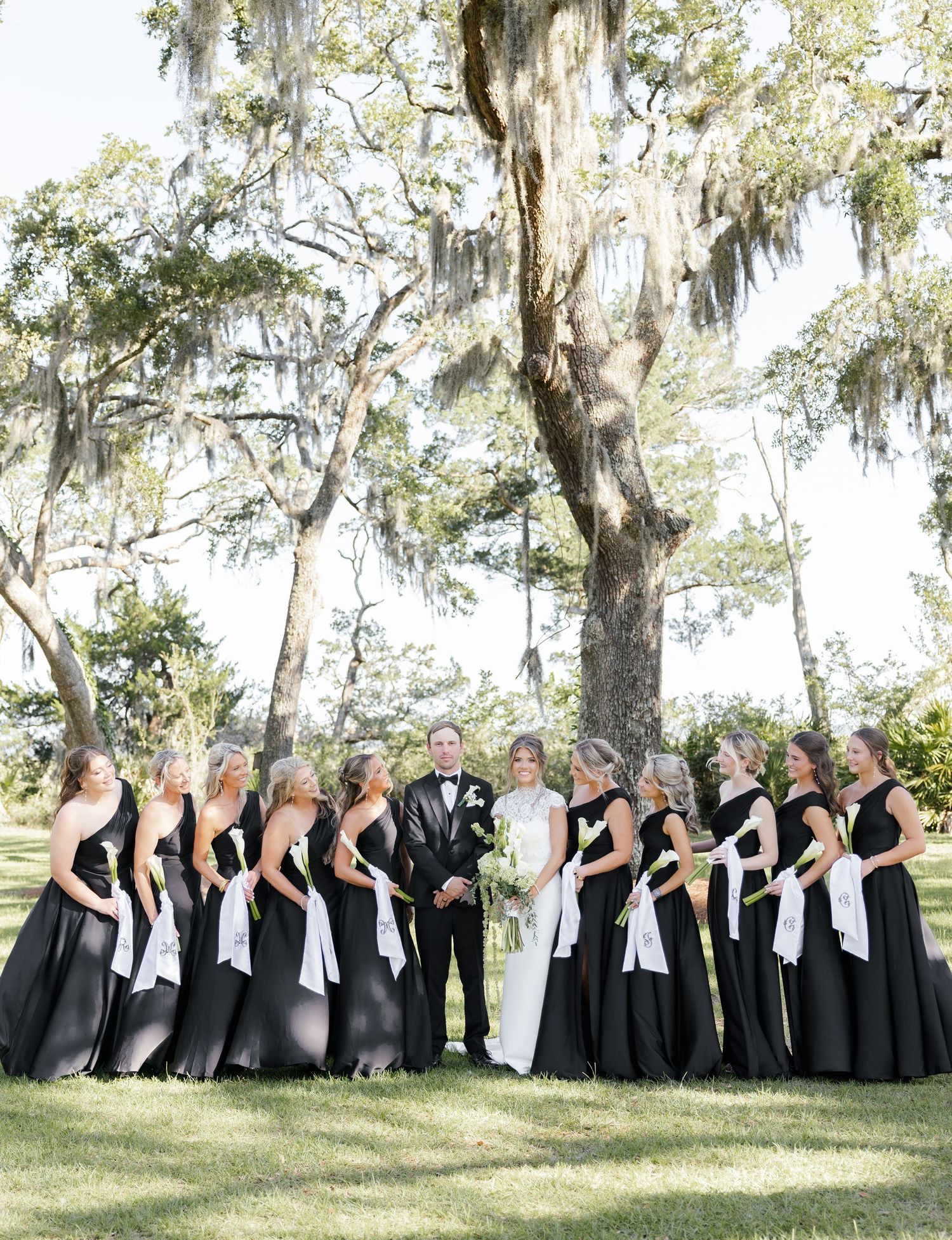 Wedding party poses outdoors with bridesmaids in black gowns with white sashes flanking bride and groom under moss-draped oak trees.