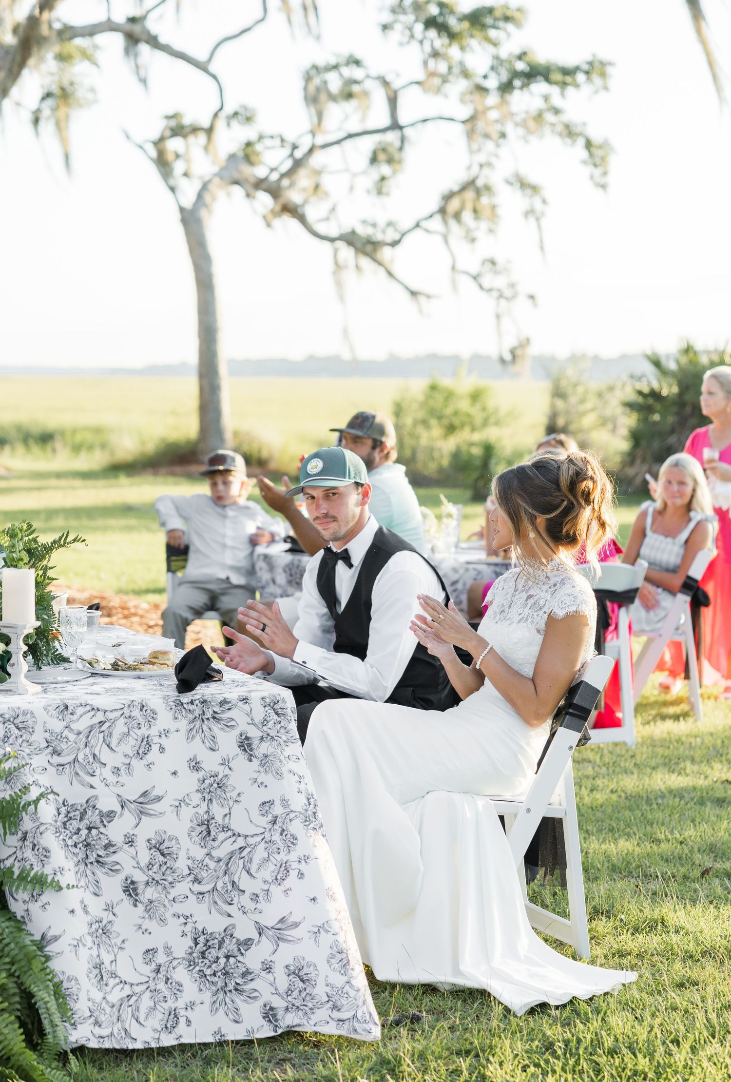 Bride and groom seated at floral tablecloth during outdoor wedding reception on lush green lawn.