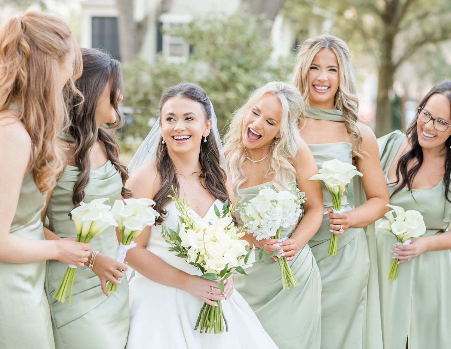 Wedding party in mint green dresses holding white bouquets pose together outdoors on sunny day.