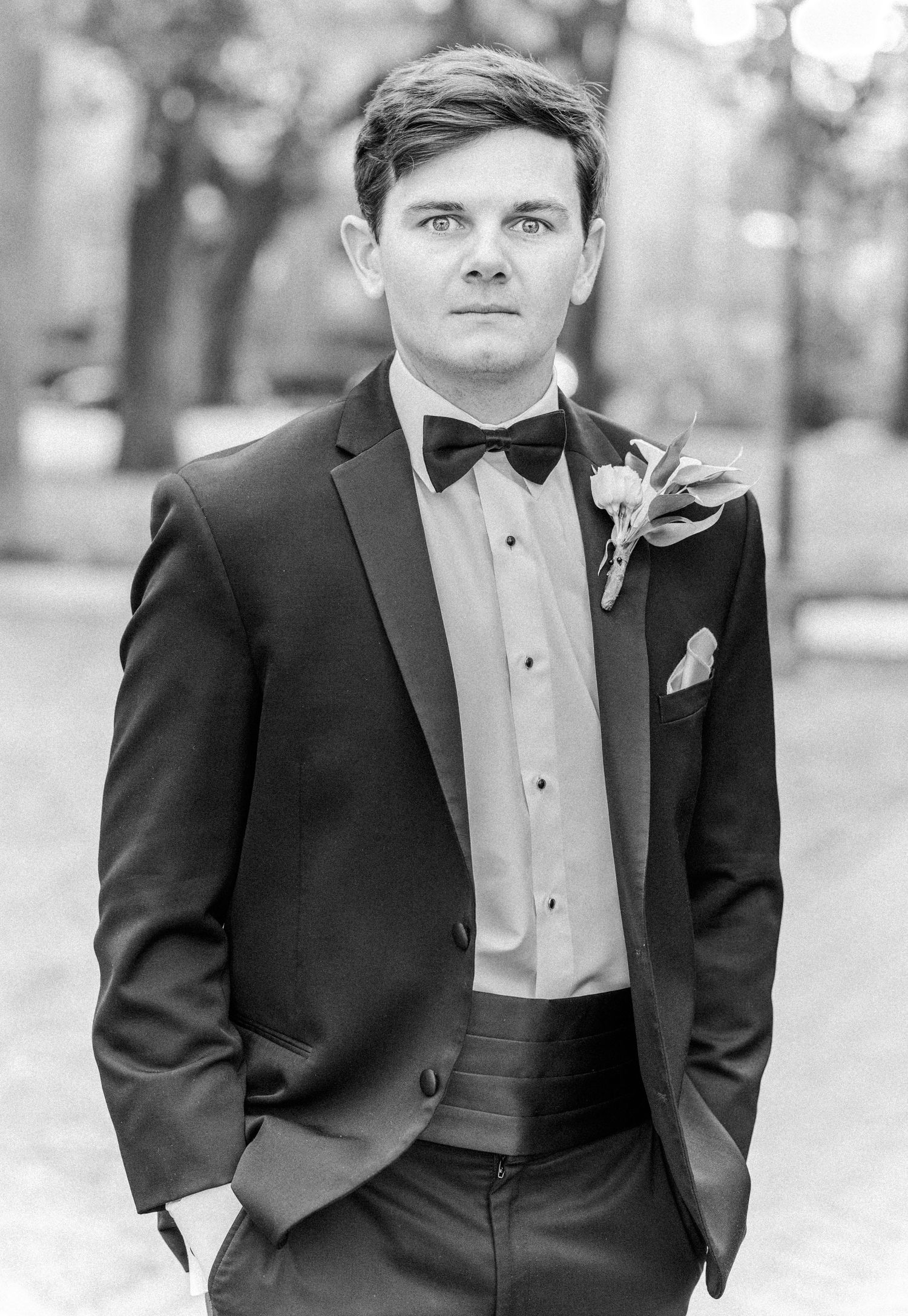 Black and white portrait of a groom in formal tuxedo with bow tie posing outdoors at a wedding.
