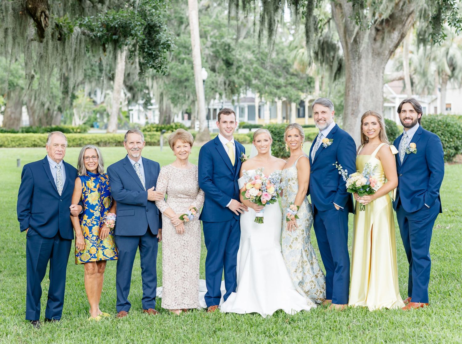 Wedding party in formal attire poses outdoors under Spanish moss draped trees in a panoramic photo.