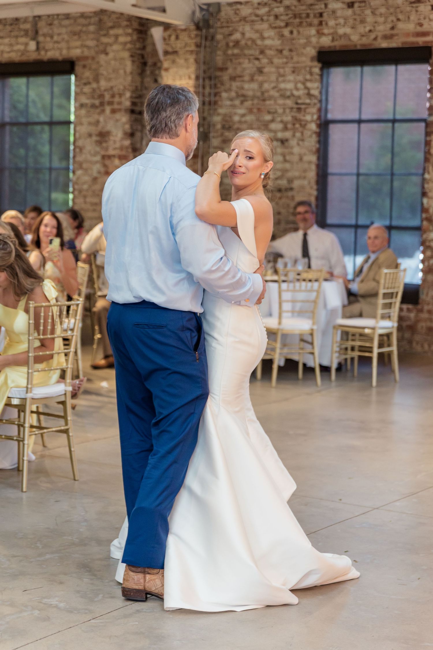 A couple dances together at their wedding reception in an industrial-style venue with exposed brick walls and chiavari chairs.