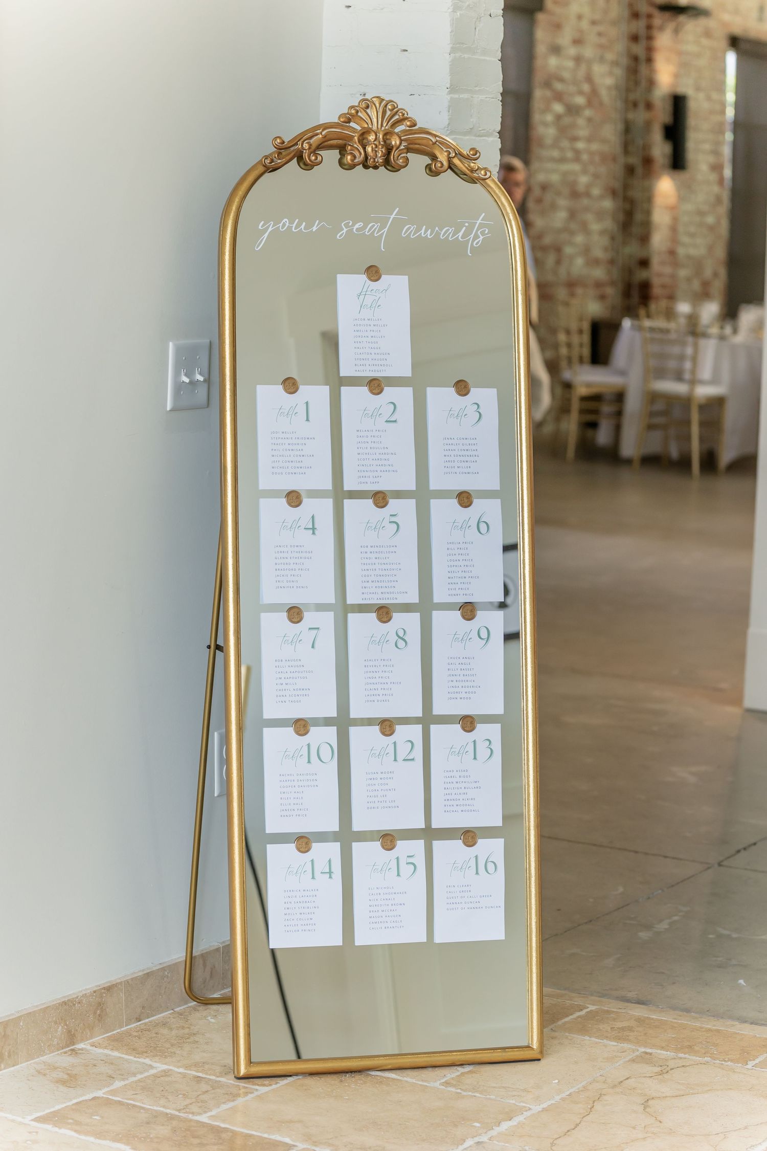 Elegant gold-framed mirror displays wedding seating chart cards arranged in rows against a rustic brick wall.