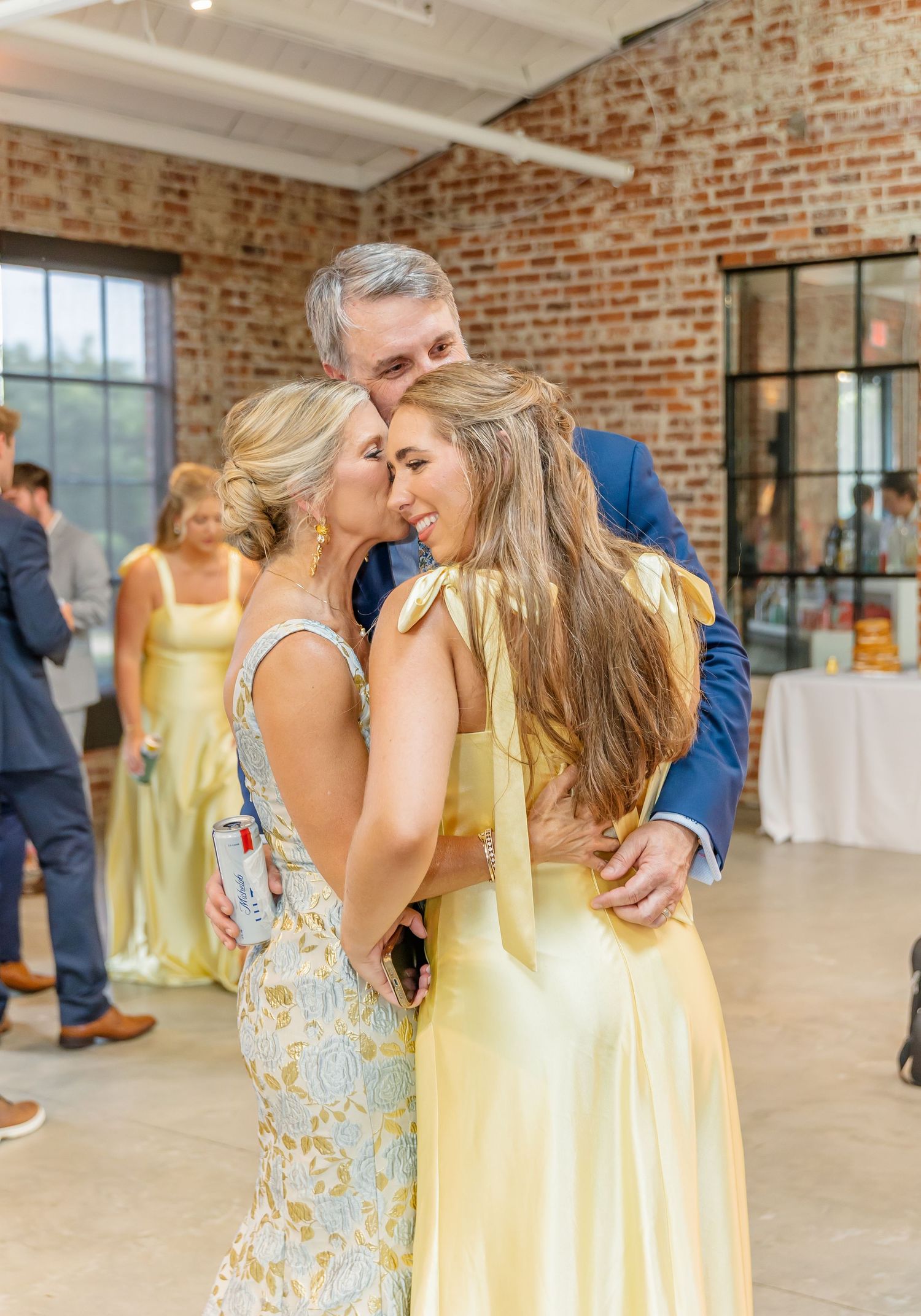 Bridesmaids in yellow dresses embrace in an industrial-style venue with exposed brick walls and large windows.