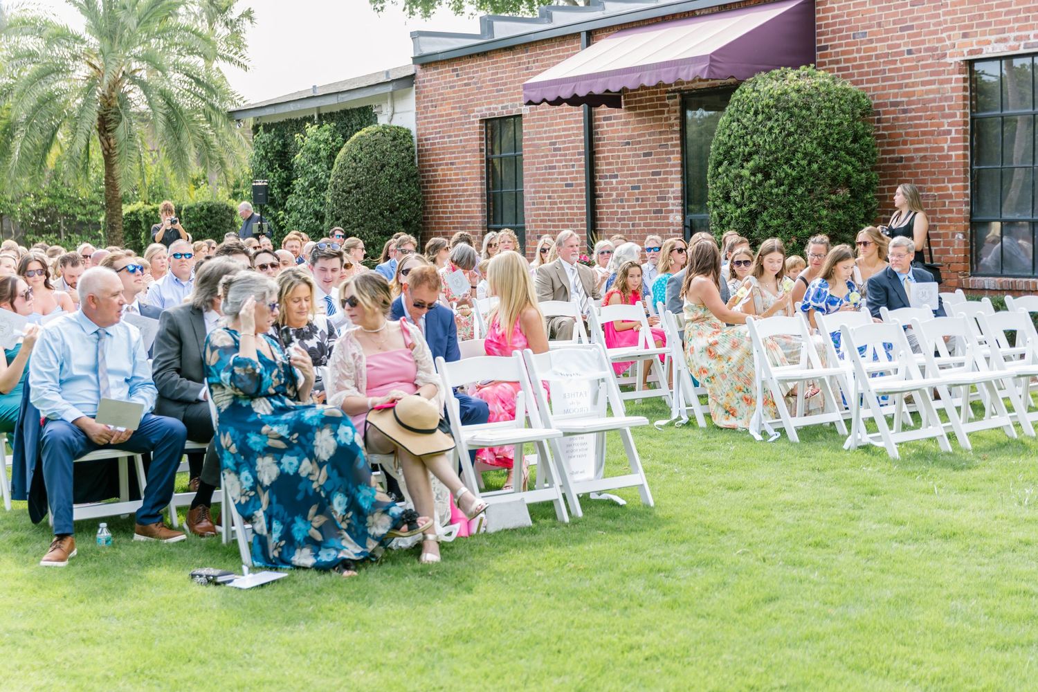 Wedding guests seated in white chairs on a lush green lawn during an outdoor ceremony at a brick venue.