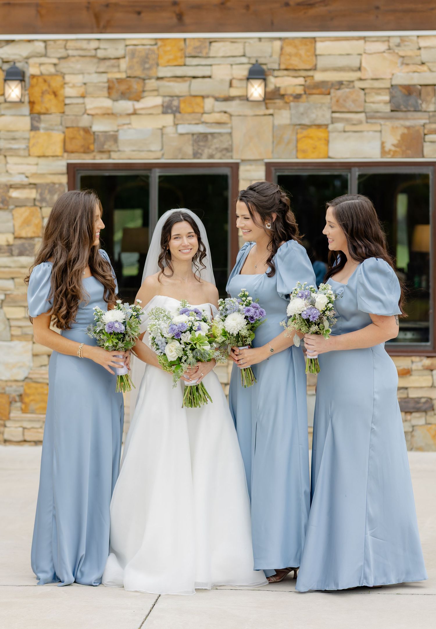 A wedding party in light blue bridesmaid dresses and white bridal gown pose together against a stone wall backdrop.