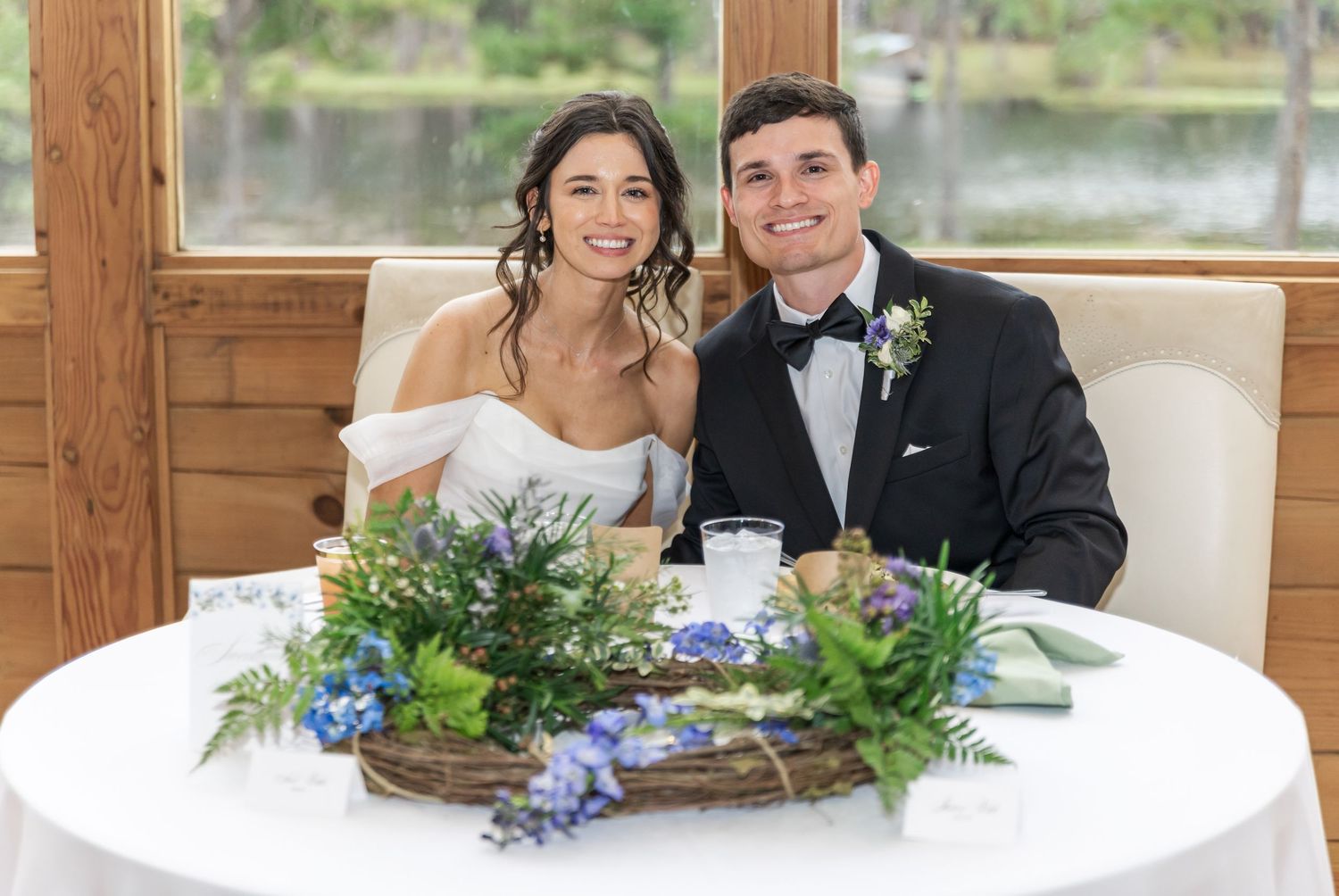 A couple sits together at a white table decorated with blue and green floral centerpieces at an indoor venue.