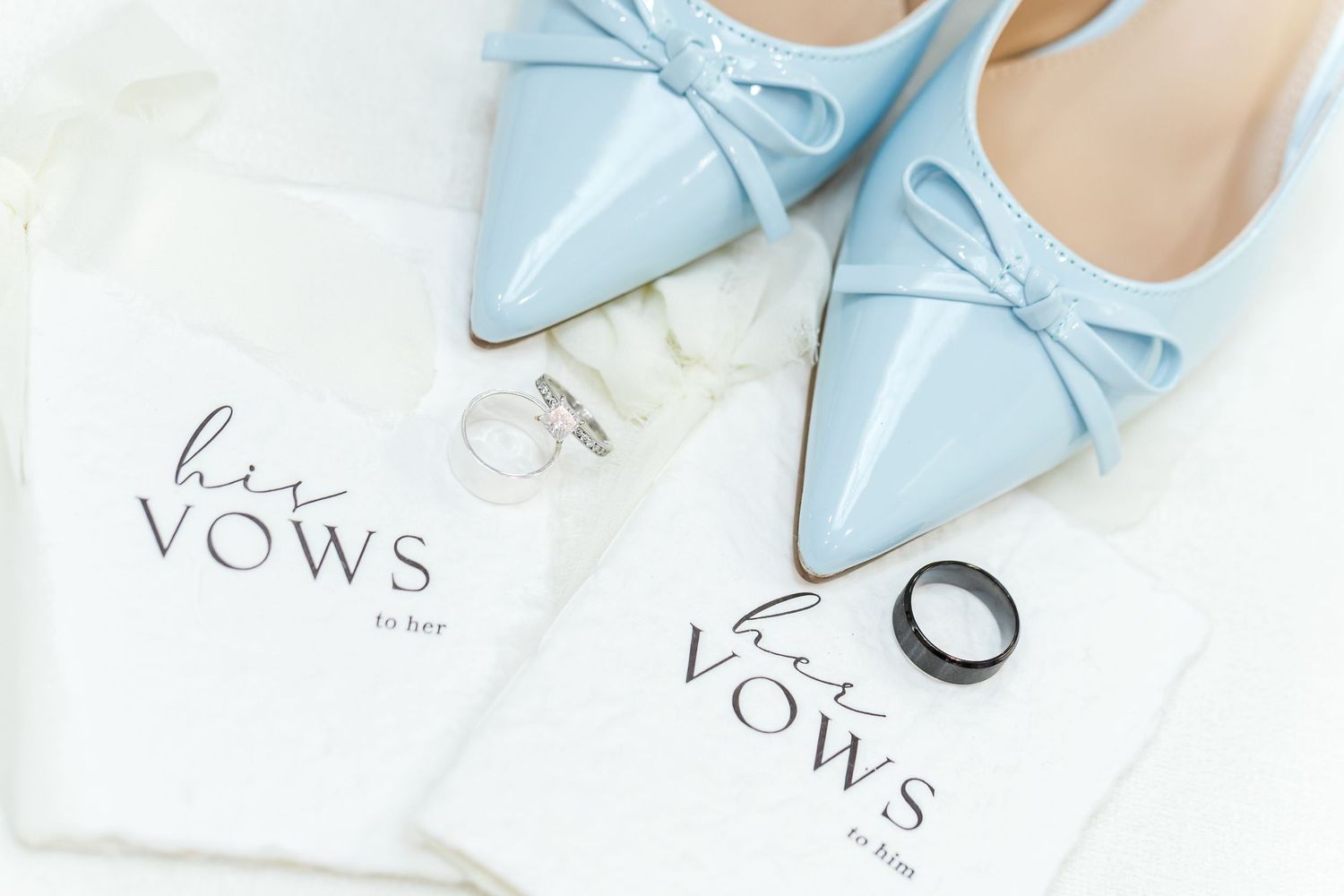 Light blue bridal heels and wedding vow books arranged on white surface with delicate styling elements.