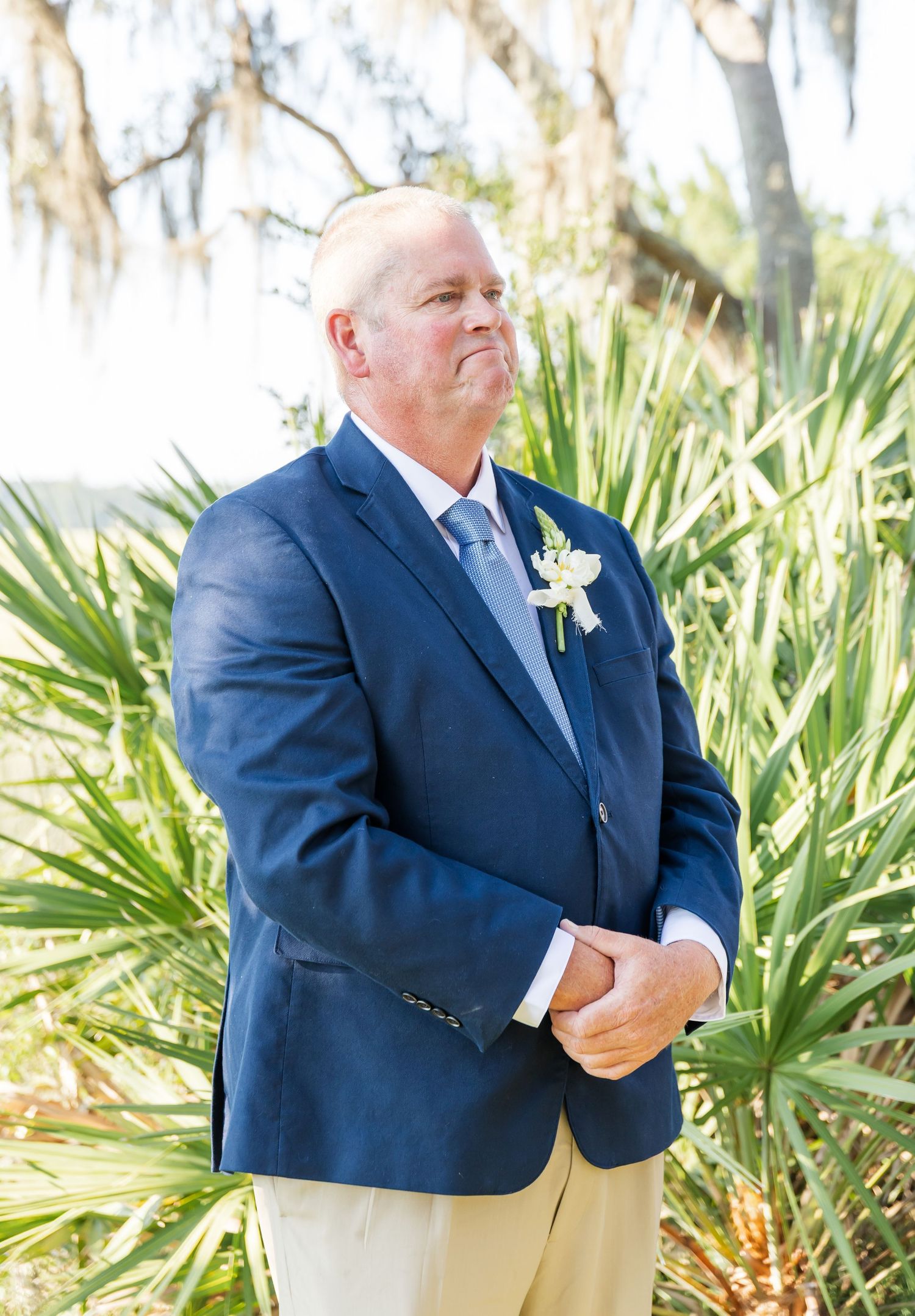 A person in a navy suit stands outdoors near tropical plants and palm fronds in bright sunlight.