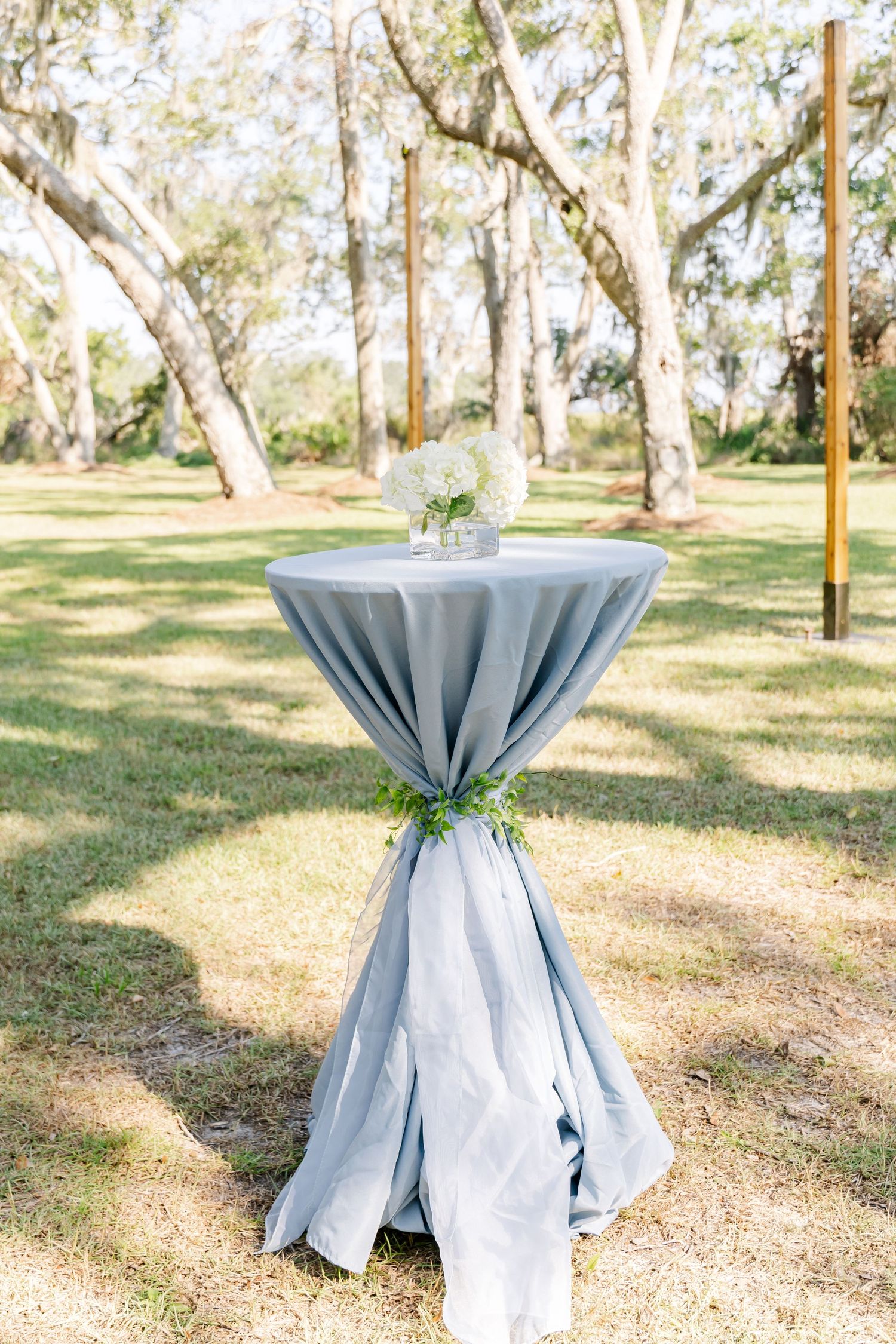 Outdoor cocktail tables with light blue tablecloths arranged in a sunny wooded area for an event.