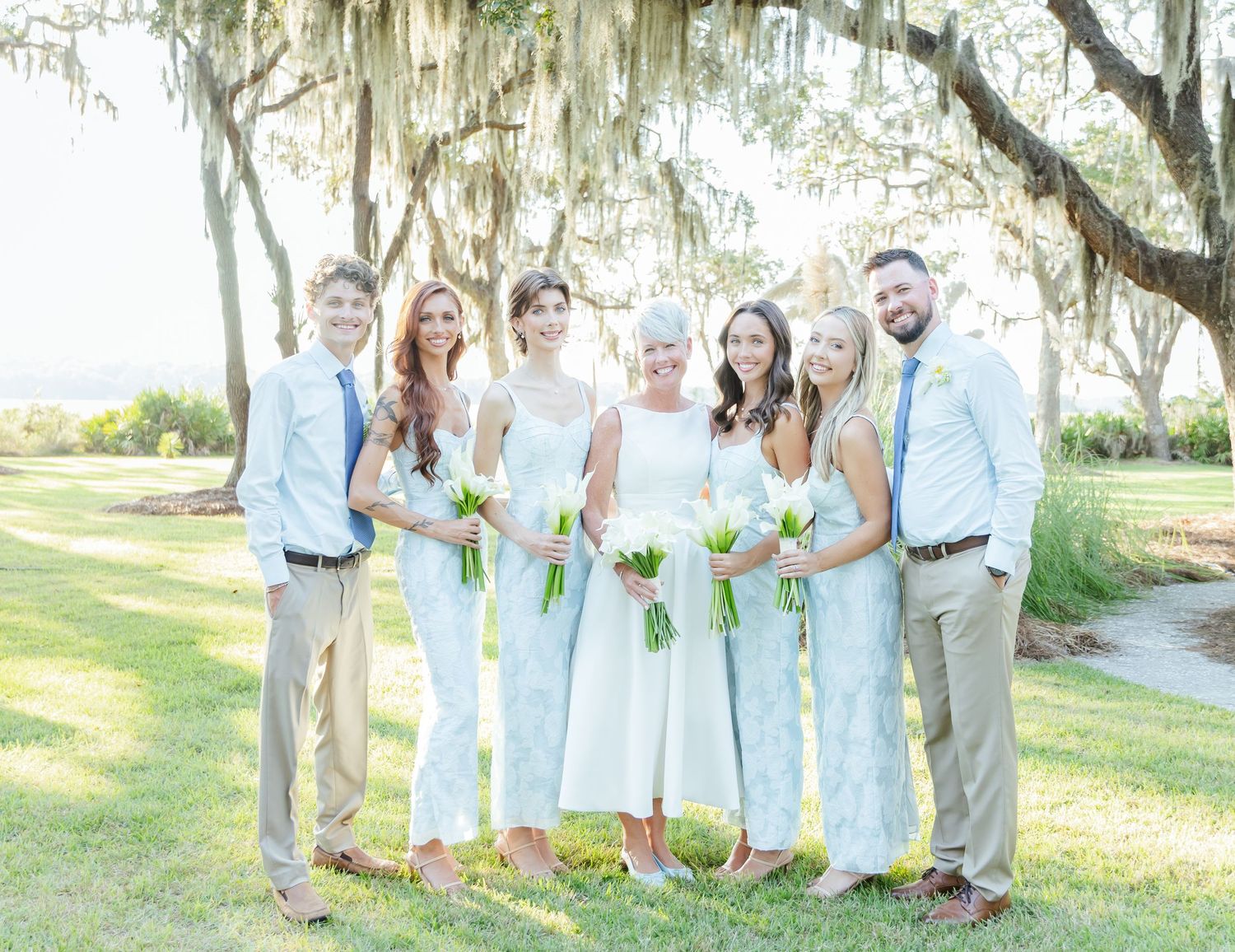 Wedding party in light blue and white attire poses together under Spanish moss draped trees at outdoor venue.