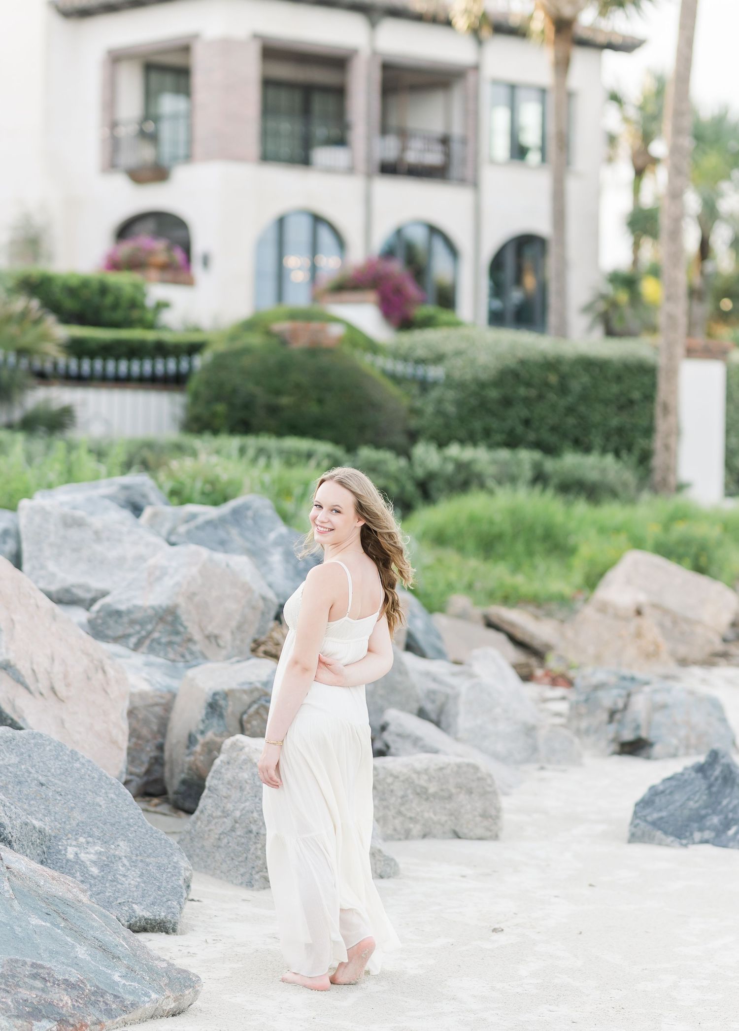 A flowing white dress contrasts against rocky coastal landscape with Mediterranean architecture in background.