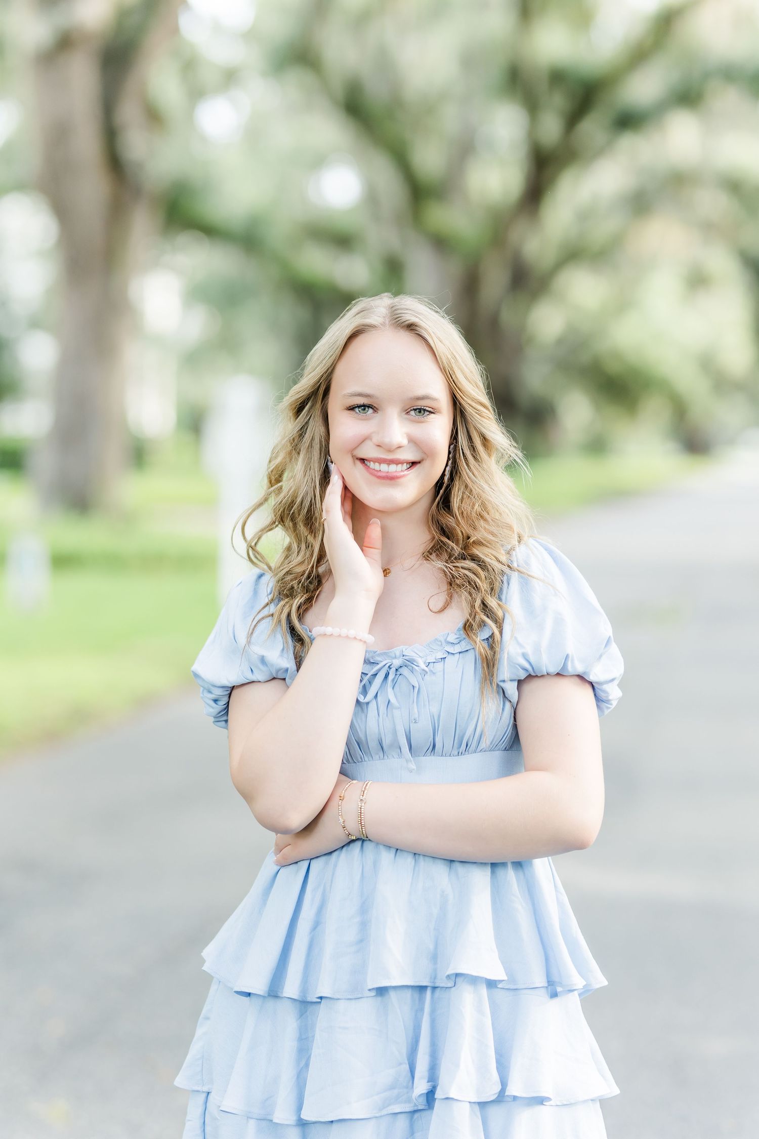 Young woman in light blue tiered ruffle dress poses outdoors on a tree-lined path.