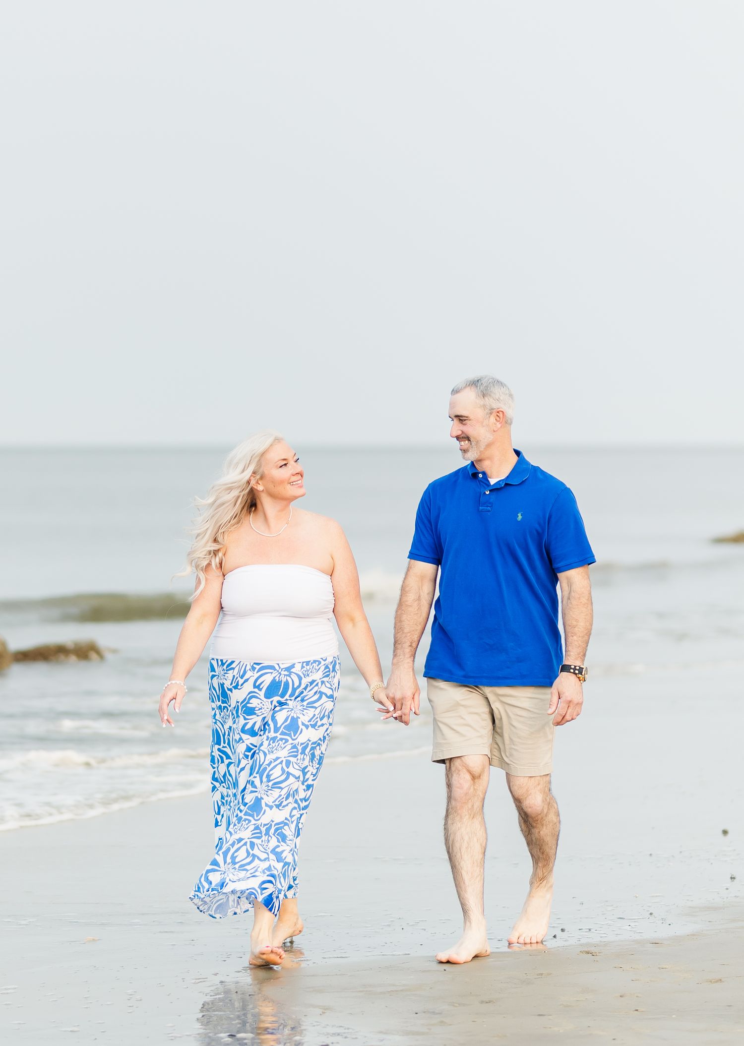 Couple holding hands and walking together on a sandy beach wearing coordinating blue and white outfits.