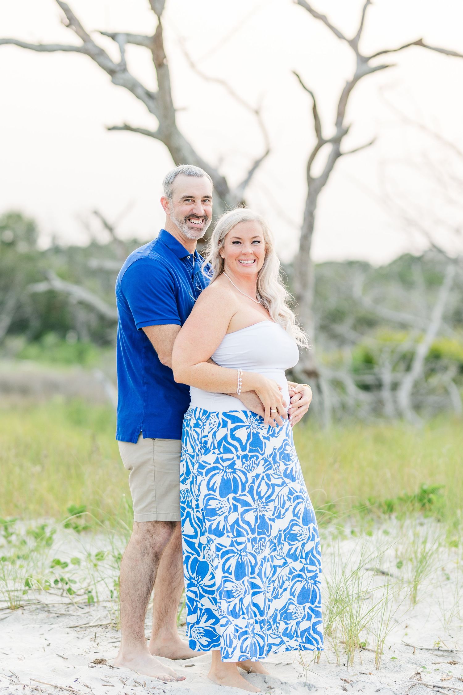 A couple embraces on a sandy beach with driftwood and vegetation in the background.