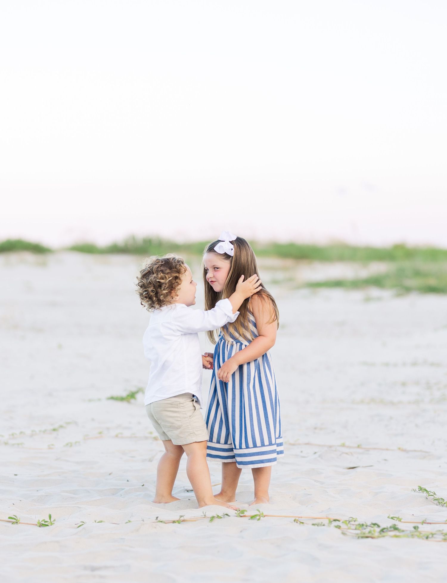 Two young children in summer outfits embrace while playing on a sandy beach.