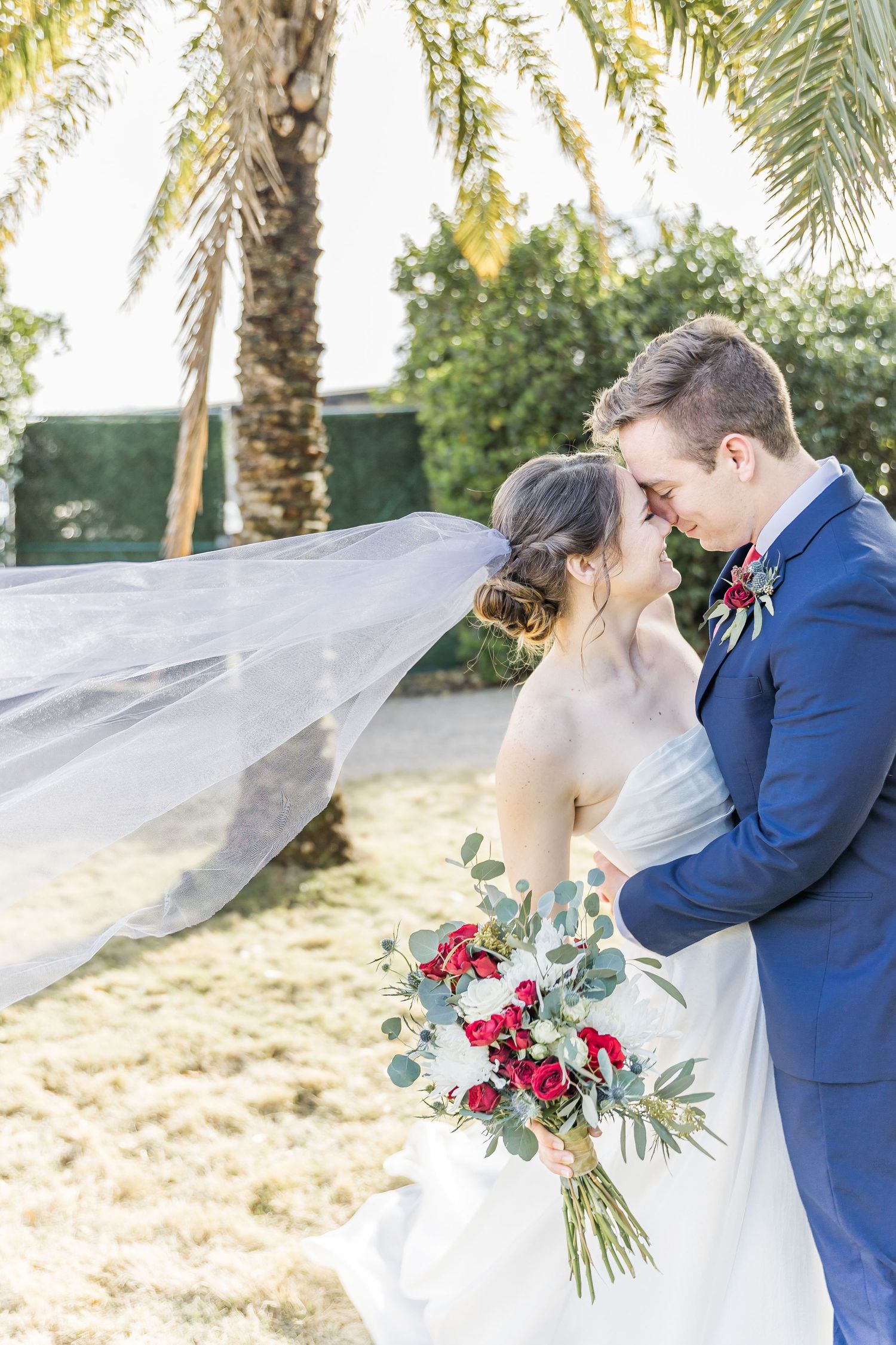 Bride with flowing veil and red bouquet shares intimate moment with groom under palm trees.