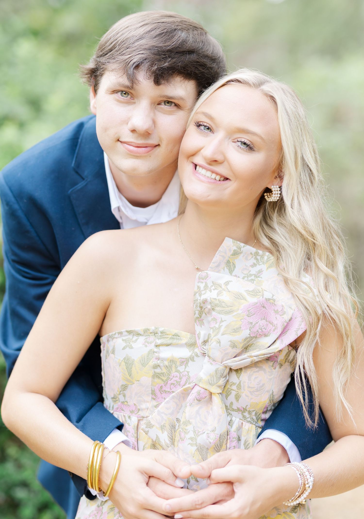 A couple smiles and embraces while wearing formal attire in an outdoor garden setting.