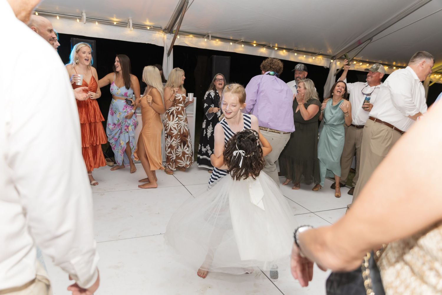 A group of people dancing at a wedding reception in a white tent with string lights overhead.
