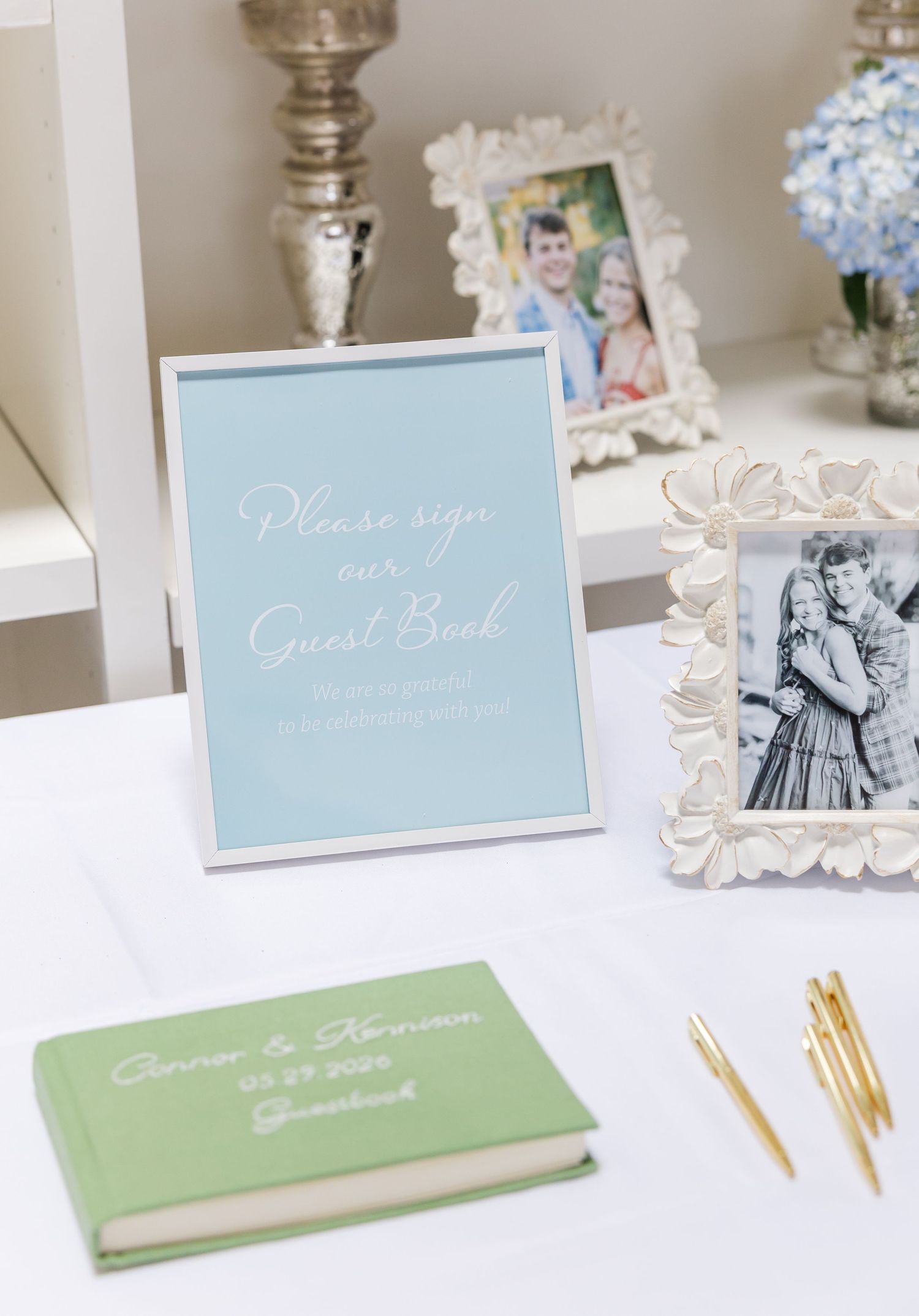 Elegant wedding guest book table with mint green book, blue sign, gold pens, and framed couple photos.