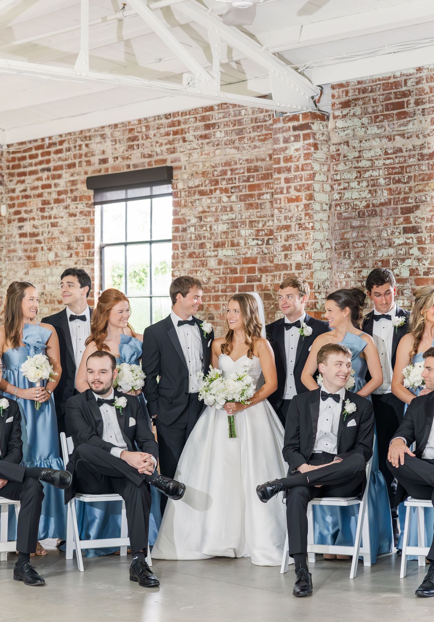 Wedding party in black tuxedos and blue gowns gathered in rustic brick venue.