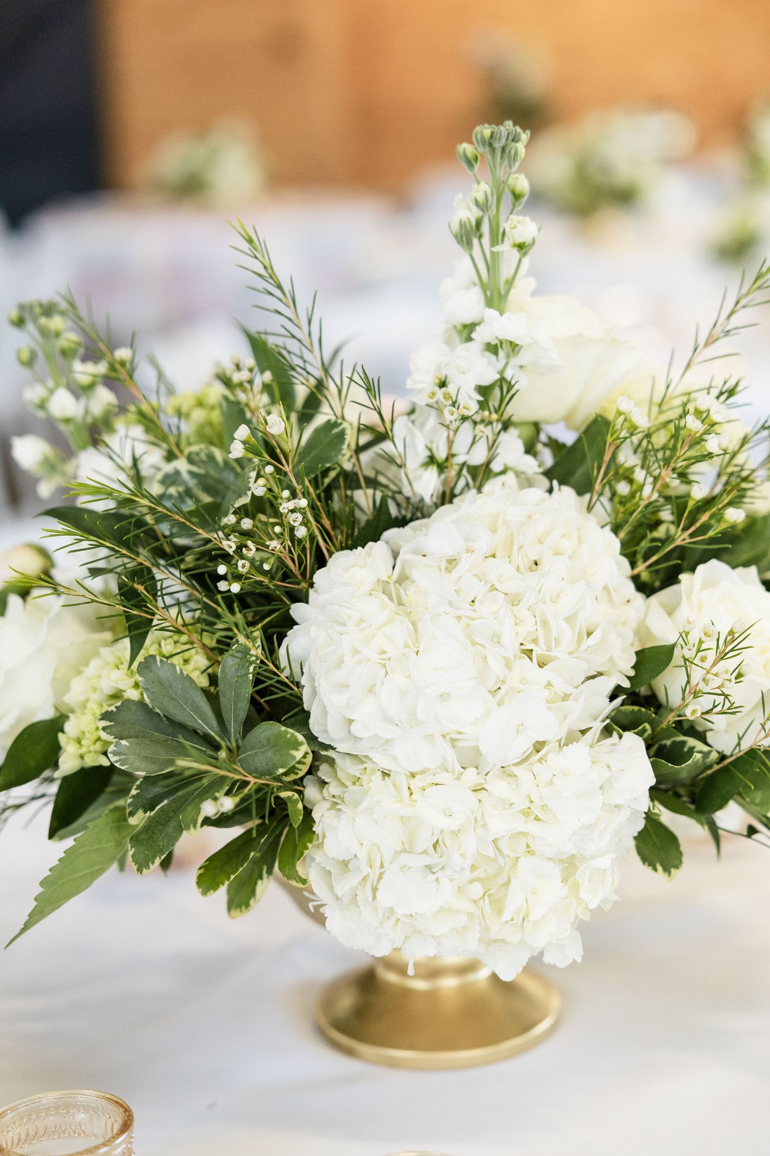 White hydrangea wedding centerpiece in gold vase with greenery on white tablecloth.
