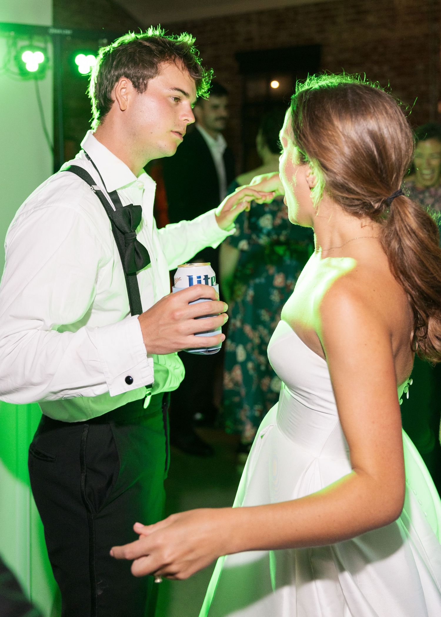 Groom holding beer can dancing with bride under green lights at lively wedding reception.