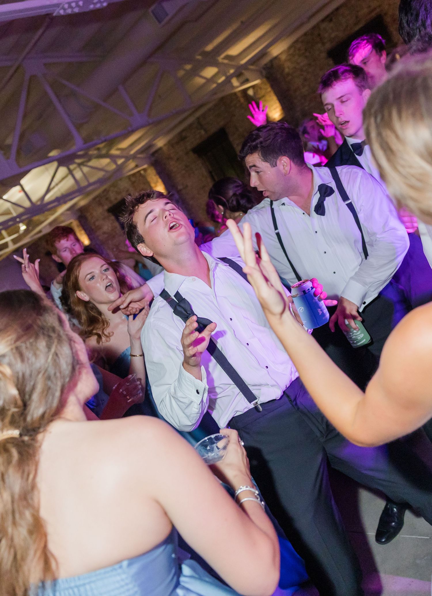 Energetic wedding reception dance floor scene with guests in suspenders dancing under purple lights.
