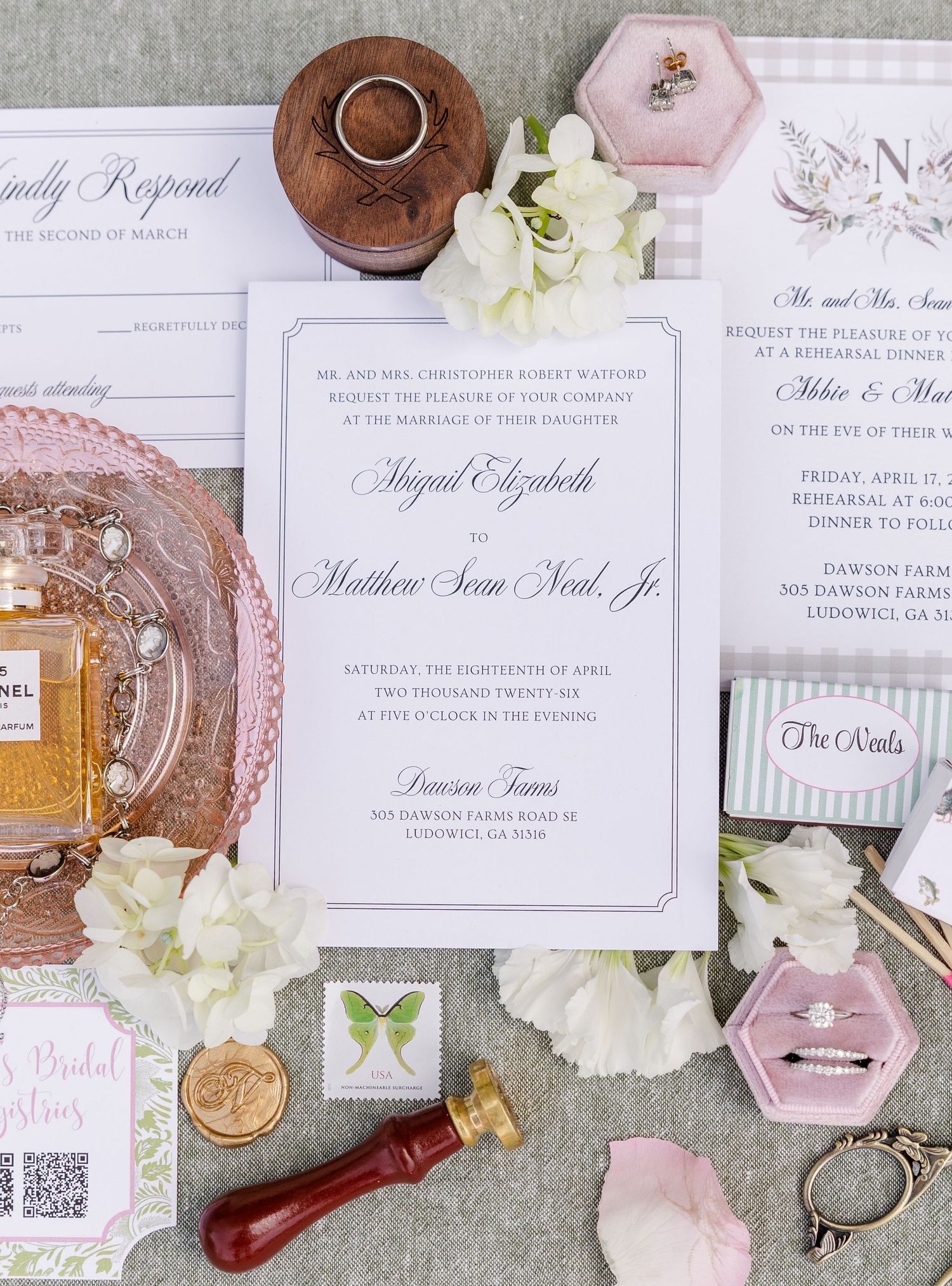 Elegant wedding invitation suite with floral accents, wax seal, rings, and pink velvet box on a soft gray background.