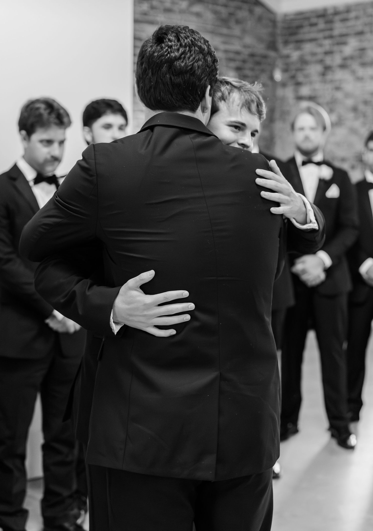 Groom sharing emotional hug with groomsman in tuxedos before wedding ceremony in black and white photo.