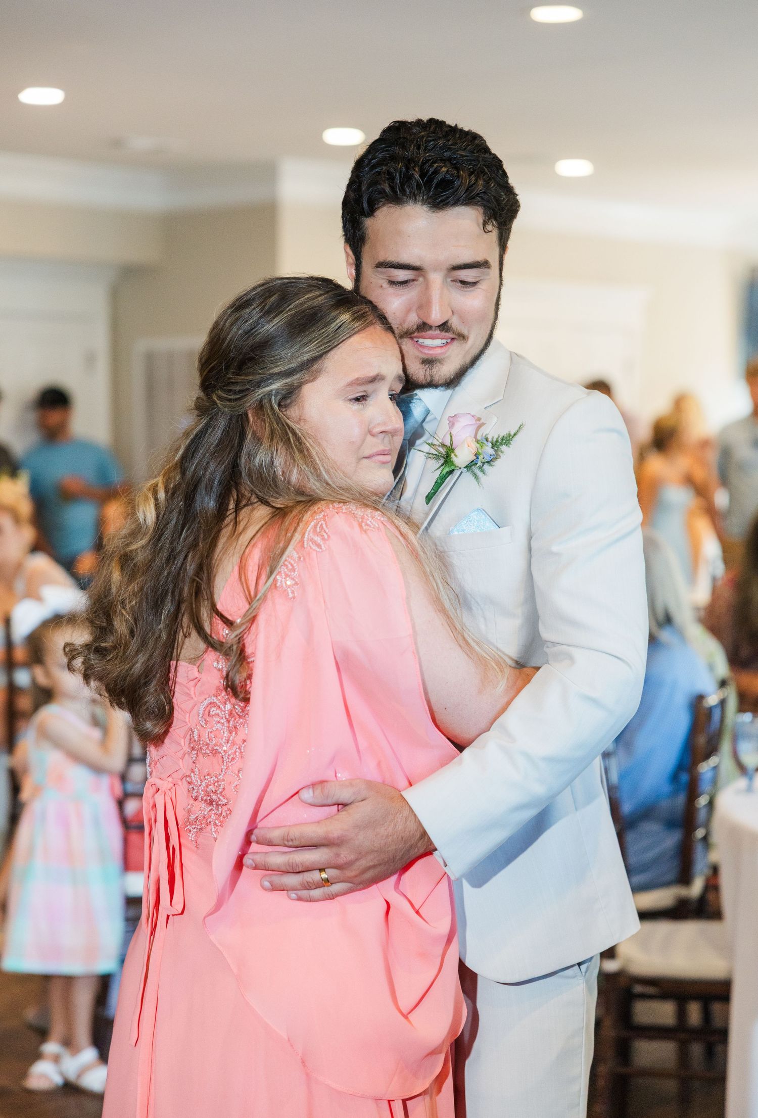 A groom in a white suit shares a tender embrace with a young girl in a coral dress at a wedding reception.