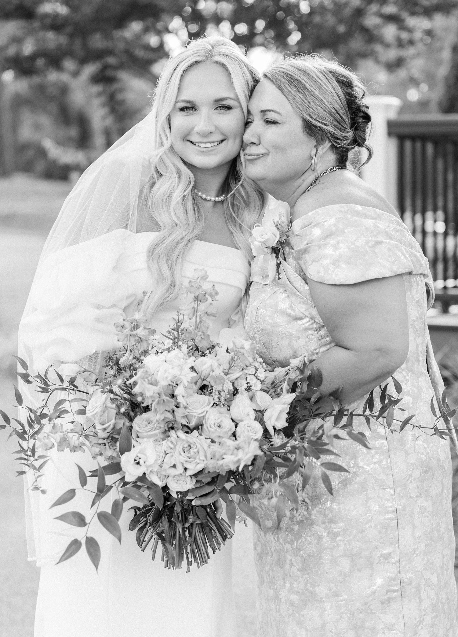 Black and white photo of smiling bride in veil holding bouquet as a woman in lace dress kisses her cheek.